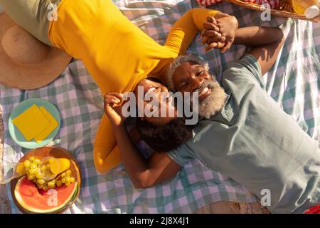 Overhead view of happy african american couple holding hands by food while lying on blanket at beach Stock Photo