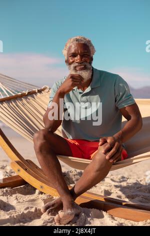 Portrait of bearded african american senior man sitting on hammock against blue sky in summer Stock Photo