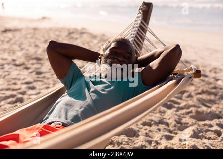 Bearded african american senior man with hands behind head lying on hammock at beach in summer
