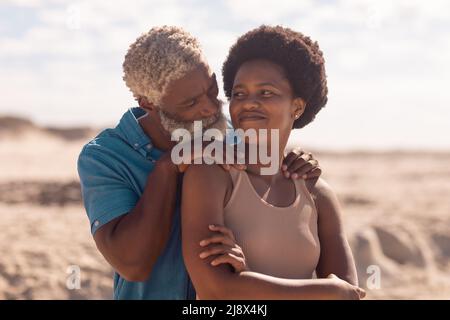 African american bearded senior man with hands on mature woman's shoulders at beach against sky Stock Photo