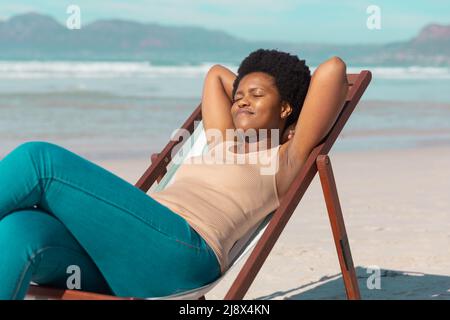 African american mature woman with hands behind head and eyes closed relaxing on deckchair at beach Stock Photo