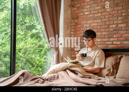 A man with cup of hot drink coffee reading book in the bedroom at home. Stock Photo