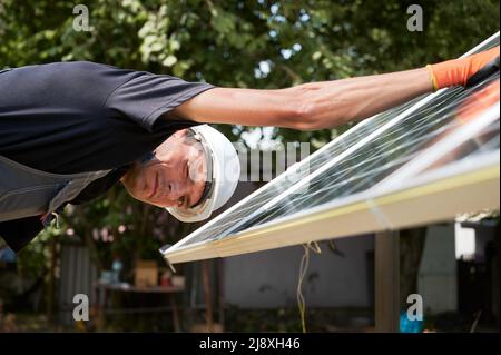 Male worker in safety helmet looking at camera and winking while mounting photovoltaic solar panel. Man assembling solar modules for generating electricity through photovoltaic effect. Stock Photo
