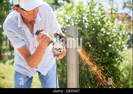 Male worker in shirt cutting metal pole with electric grinder. Man in protective glasses using hand-held circular saw while preparing aluminum pole for solar panel installation. Stock Photo