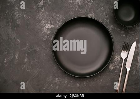 Black ceramic plate mockup with black sauce bowl and cutlery set on stylish dark background. overhead shot Stock Photo