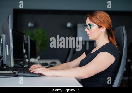 Redhead woman in glasses and casual wear working overtime. Focused girl alone works on a computer in an empty office. Deadline. The IT specialist Stock Photo