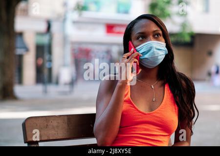 Woman wearing a face mask while talking on the phone sitting on a bench outdoors. Stock Photo