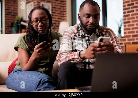 Boyfriend ignoring upset girlfriend after arguement spending time on smartphones sitting on couch. African american couple living together having issues not talking to each other in home living room. Stock Photo