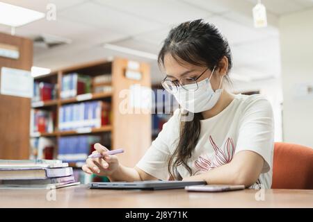 In the library, an Asian woman searches for information using a tablet and a book. Stock Photo