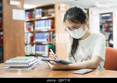 In the library, an Asian woman searches for information using a tablet and a book. Stock Photo