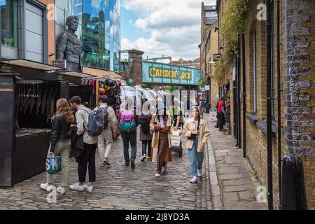 A busy street filled with shoppers and tourists, with the iconic Camden Lock railway bridge in the background. Stock Photo
