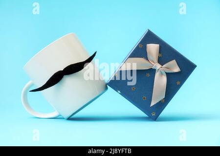 Close up of white mug with a felt moustache and present box on a blue background. Concept of Father's day. Stock Photo