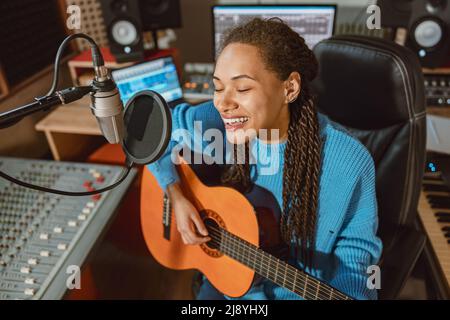 Enthusiastic multi-ethnic woman singer plays guitar and recordis song, sings into microphone in music recording studio Stock Photo
