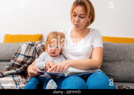 Portrait of young mother is reading a book to her child sitting on the sofa. Preschool education. Stock Photo