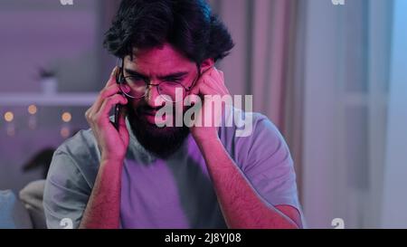 Arabic Indian bearded man with glasses talking on phone at house party sitting on sofa neon light hard to hear conversation loud music noisy birthday Stock Photo