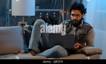 Freelancer guy Arab Indian man bearded male user wearing glasses sitting on sofa at night late time with laptop reading news online thinking doubts jo Stock Photo