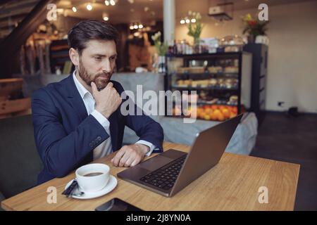 Handsome Caucasian thoughtful serious businessman sitting at laptop and thinking in cafe. Stock Photo