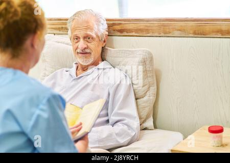 Nurse reading from a book by the bedside of a sick senior in a nursing home Stock Photo