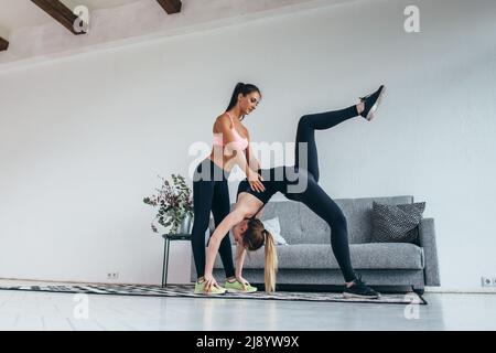 Fitness woman doing advanced bridge pose called one-legged wheel with assistance of instructor at home Stock Photo