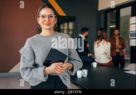 Modern businesswoman looking at the camera while holding a digital tablet. Cheerful young businesswoman standing in a boardroom with her colleagues in Stock Photo