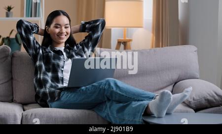 Asian woman Korean girl freelance student lady user sitting on sofa with hands behind head resting work break stop e-learning watching movie online vi Stock Photo