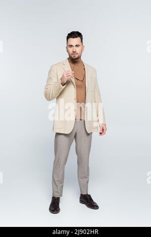 full length of critic in beige blazer and trousers holding eyeglasses on grey Stock Photo