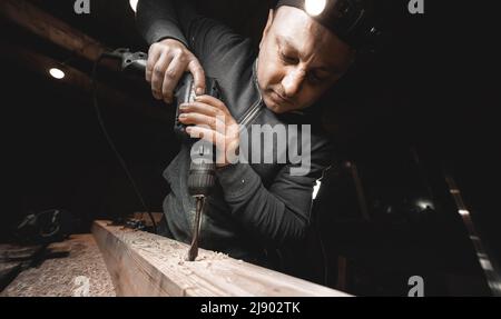 A man works with a drill in his workshop. Carpenter drills with a hand power tool in a dark room with directional light Stock Photo