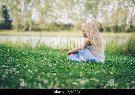 adorable girl with long blond hair sitting in green clover floral meadow Stock Photo