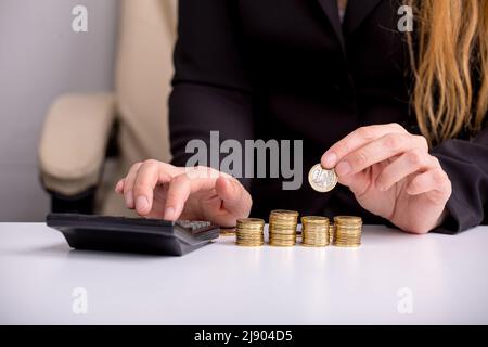 The girl is counting on a calculator at the table, in front of stacks of coins, close-up Stock Photo