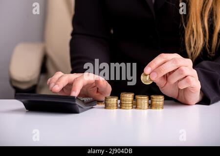 A business woman counts on a calculator and holds a coin in her hand, at a table, in front of stacks of coins, close-up Stock Photo
