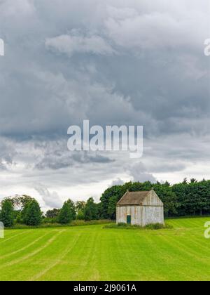 A pale white Old Doocot or Dovecote situated in a field of newly sown wheat with a mixed woodland behind and dark clouds above. Stock Photo