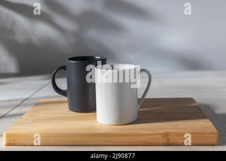 Two coffee cups - white and black on a wooden plank on the table. On the wall, the shadow from the trees from the window Stock Photo
