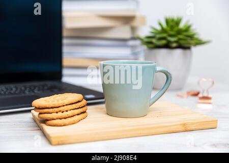 Blue cup and cereal cookies on a wooden board. Blurred computer and stack of books in the background Stock Photo