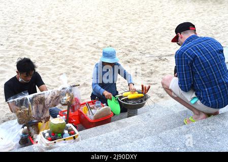Nha Trang, Vietnam - 17 May 2022: Young man orders a grilled maize from vietnamese people at beach of Nha Trang Stock Photo