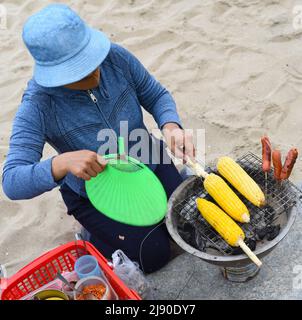 Nha Trang, Vietnam - 17 May 2022: Vietnamese woman cooks grilled maize at beach of Nha Trang Stock Photo