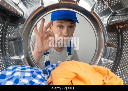 Male repairman in special clothes shows an OK sign with his hand, photo from inside the drum of the washing machine. Concept of repair work. Stock Photo