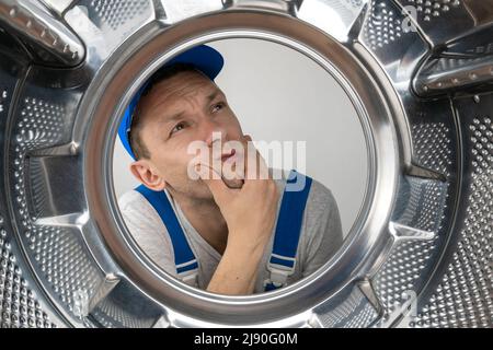 Male repairman in uniform holds his chin and looks thoughtfully to the side, photo from inside the drum of a washing machine Stock Photo