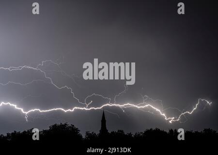 Rain falling and stroke of forked lightning during thunderstorm at night over church tower and trees Stock Photo