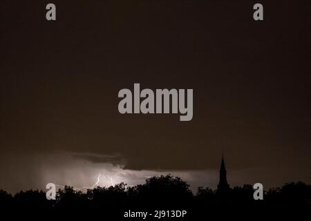 Rain falling and stroke of forked lightning during thunderstorm at night over church tower and trees Stock Photo