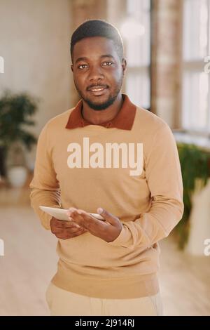 Portrait of smiling handsome young bearded marketing specialist in beige sweater using tablet in loft office Stock Photo