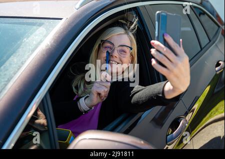 middle-aged blonde woman in glasses making selfie with a smartphone through an open car window Stock Photo