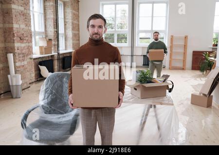 Portrait of content handsome young office man in turtleneck holding closed moving box against desk covered with polyethylene Stock Photo