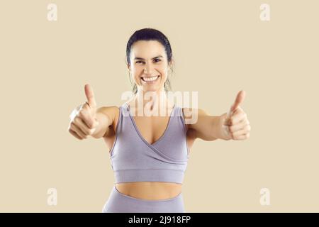 Beautiful happy sports woman in sportswear showing thumbs up on beige background. Stock Photo