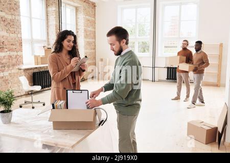 Smiling young woman checking list while her colleague unpacking moving box of documents in new office Stock Photo