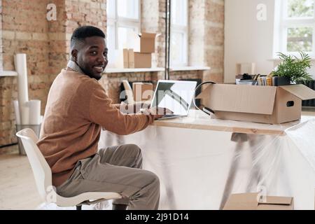 Portrait of cheerful young African-American employee in casual sweater sitting at desk with moving box and typing on laptop in new office Stock Photo