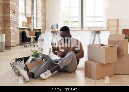 Young Afro-American man sitting among cardboard boxes and using laptop while eating noodles in new office after moving Stock Photo