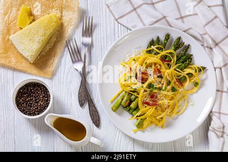 roasted asparagus spears with spaghetti sprinkled with shredded cheese, fried bacon pieces and parsley on white plate on white textured wood table, fl Stock Photo