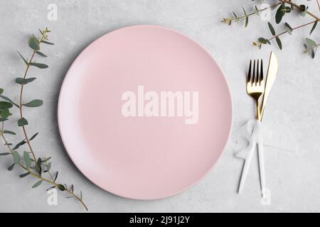 Festive table setting with pink ceramic plate, golden cutlery set and fresh eucalyptus leaves on grey concrete table top view. Plate mockup. Copy spac Stock Photo