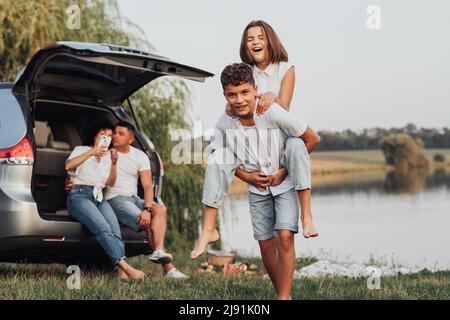 Two Happy Teenage Children Playing by the Lake While Their Parents Sitting in Trunk of the Minivan Car, Four Caucasian Members Family Enjoying Road