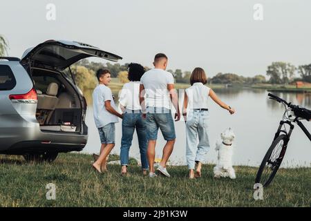 Four Members Family Traveling by Minivan Car and Having Picnic at Sunset by the Lake, Mother and Father with Two Teenage Children and Pet West Stock Photo